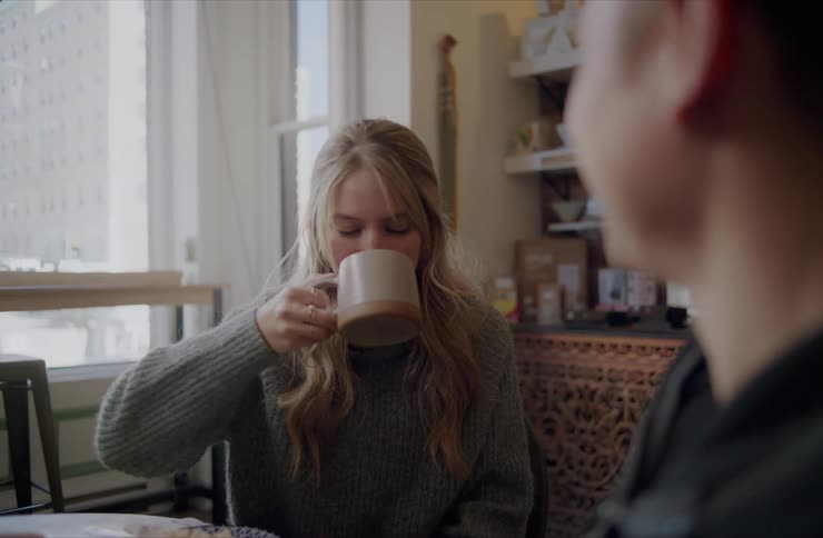 A young woman sips from a coffee mug while talking with a man over breakfast