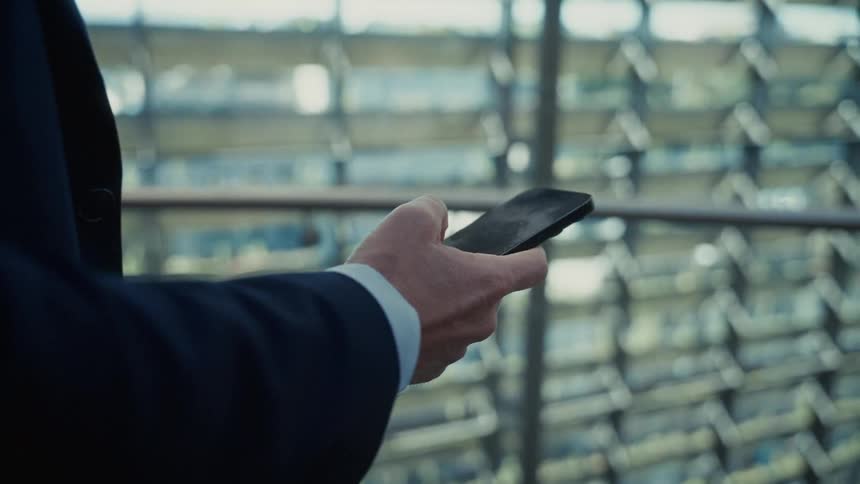 A persons hand wearing a dark suit jacket and white cuff holds and operates a mobile phone against a background of blurry windows and architectural slats