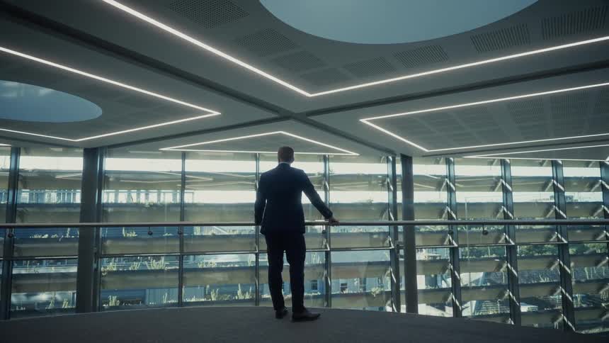 A person wearing a business jacket stands with hands resting on a metal barrier inside a contemporary office looking through a wall of slatted windows at exterior vegetation under a ceiling with geometric light patterns