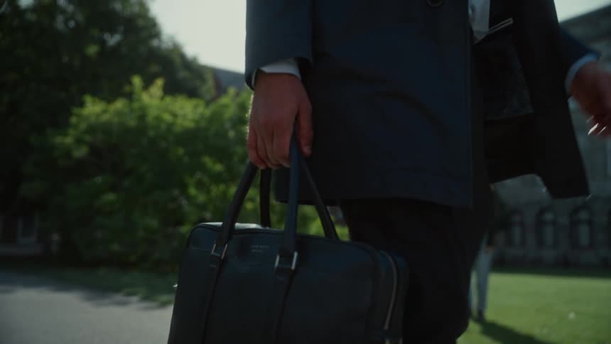 The video shows a businessperson walking outside on a sunny day holding a black briefcase or laptop bagthe person is dressed in a dark suit and the bright sunlight creates lens flare and strong shadows against the surrounding green trees and