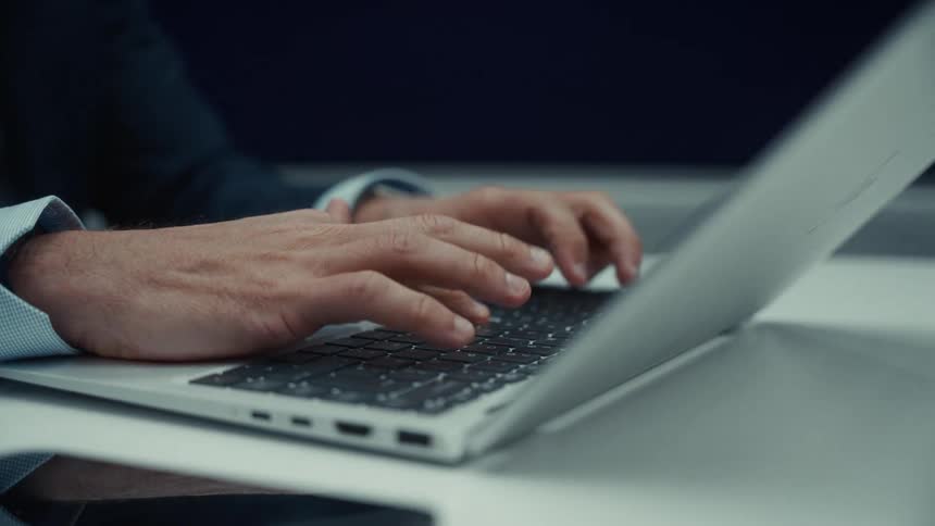 A persons hands move across the keyboard of an open portable computer the keys are pressed rapidly the laptop sits on a flat surface and the person wears a dark jacket and a collared shirt