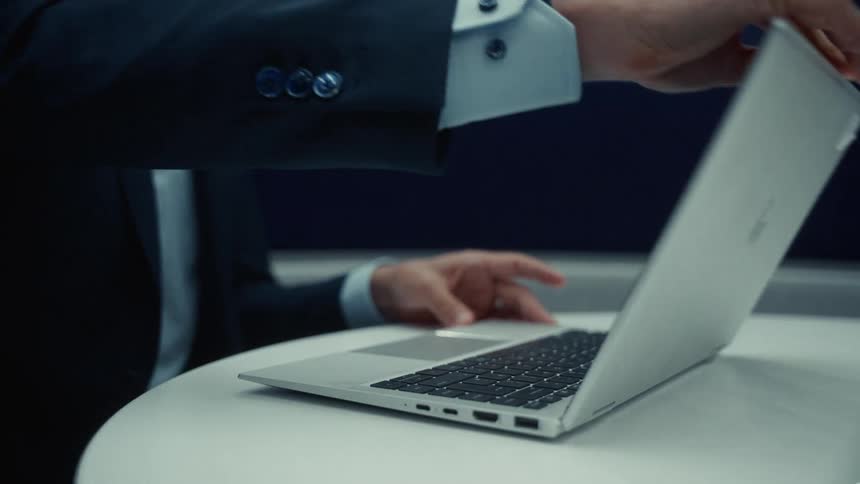 A man in a business suit places a silver laptop on a white table opens the screen and immediately begins using the laptop