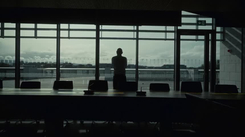The silhouette of a person stands centrally against a large glass window overlooking a city framed by a long conference table and multiple chairs in the foreground of a darkened room