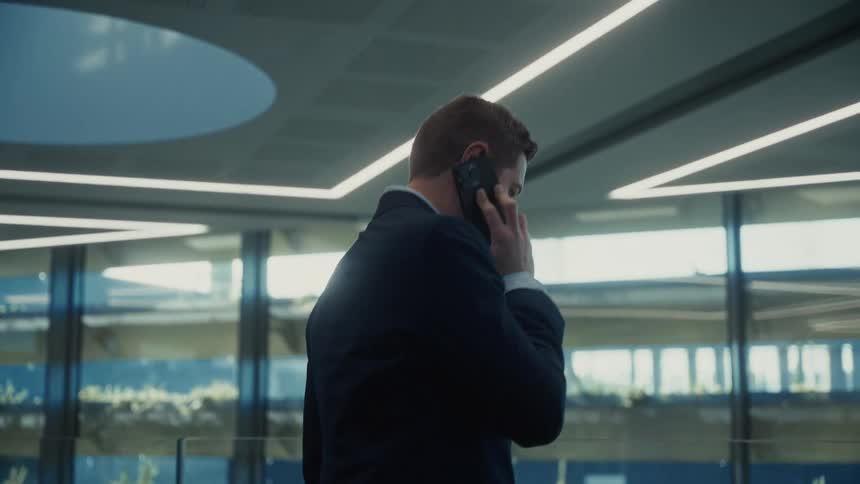 A medium rear view shows a businessman in a dark suit walking through a modern glass office building interior while talking on his mobile phone