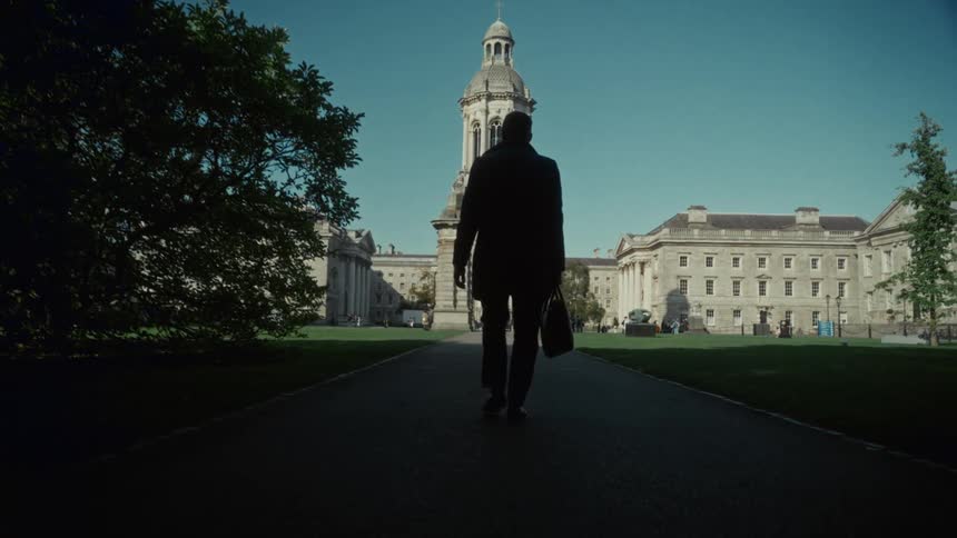 A person walks down a paved path toward a university building a central tower stands at the end of the walkway dark foliage frames the near ground and theres a clear sky