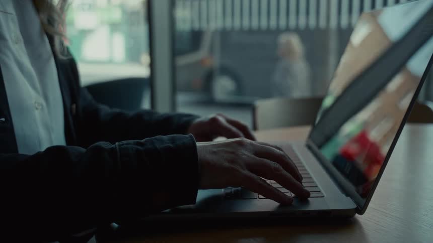 A person sits at a wooden table in an indoor space near a large window opens a silver laptop computer places both hands on the keyboard and begins typing while cars move outside on the street