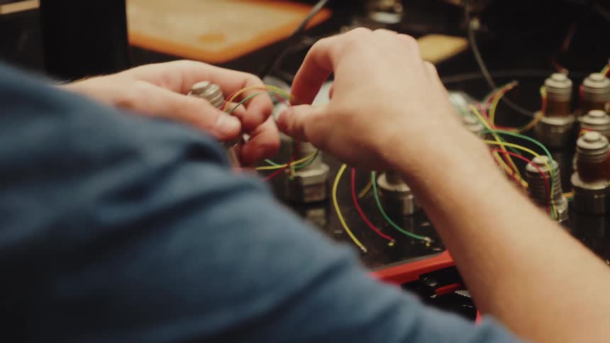 Two hands hold and manipulate thin colorful electrical wires near several metal cylindrical components while a pair of pliers and other cables rest on a dark workbench