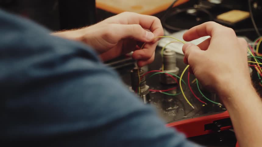 A persons hands manipulate thin colored conductors over a dark workstation the fingers strip insulation from one conductor a tool rests on the surface and several components are visible in the foreground