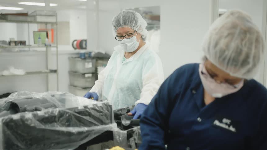 Two women wear head covers and face protection in a laboratory both women work with dark objects covered in plastic film with shelves in the background
