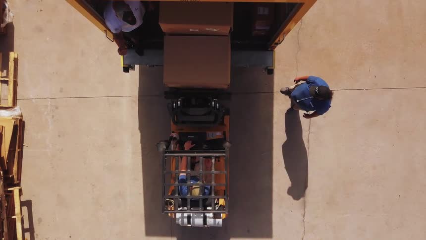 A motorized lifting vehicle carrying two brown boxes backs out of a large yellow truck trailer two workers stand nearby on a concrete surface and wood pallets are stacked on the side of the area