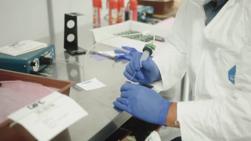 The video shows an extreme closeup of a persons hands wearing blue nitrile gloves carefully applying a liquid from a syringe to a clear reflective glass surface on a tabletop the worker is dressed in a clean room suit or white lab coat with