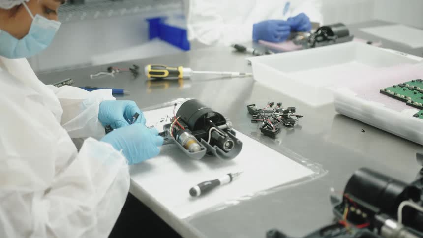 The video shows an industrial setting where a person in a cleanroom suit and face mask is assembling a device the worker is using their gloved hands to manipulate small complex mechanical parts of a black cylindershaped object