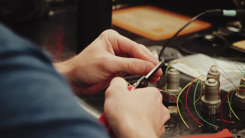 A close view shows the hands of a worker wearing a dark shirt cutting a small multicolored electrical wire with wire cutters on a workbench with electronic components