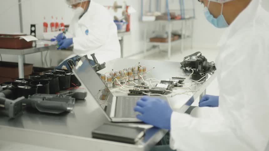 Two professionals in sterilized clothing operating electronic components on metal bench in cleanroom
