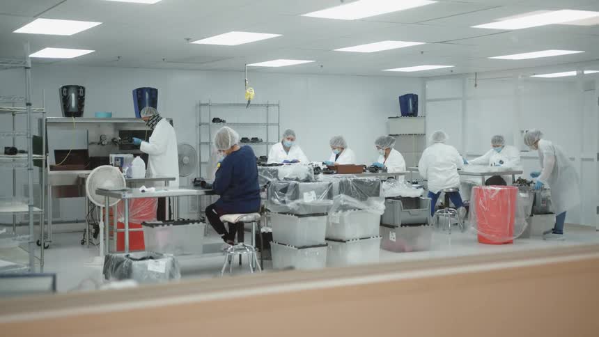 A view through an interior window shows a brightly lit clean room with a team of workers wearing white lab coats hair nets and face masks sitting at tables performing assembly work