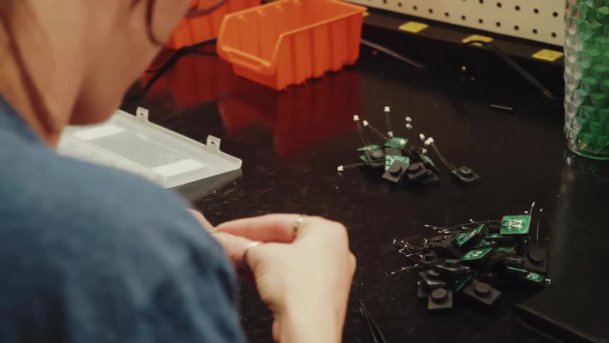 A young woman sits at a workbench manipulating wires and small electronic components with a pile of circuit boards nearby and storage containers visible in the background