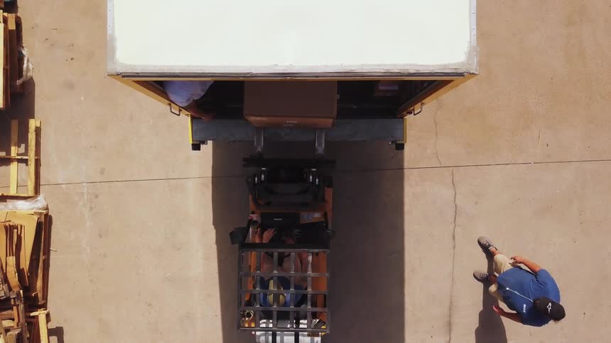 A motorized lifting vehicle moves a single large cardboard box forward into the open back of a white truck trailer while two workers stand on the concrete pavement observing the loading operation