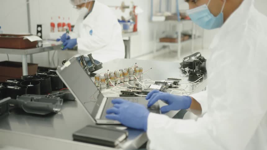 The video shows a worker in a cleanroom or laboratory setting wearing a lab coat a face mask and blue gloves they are actively working on a laptop computer that is open on a metallic countertop in the background another worker is partially vi