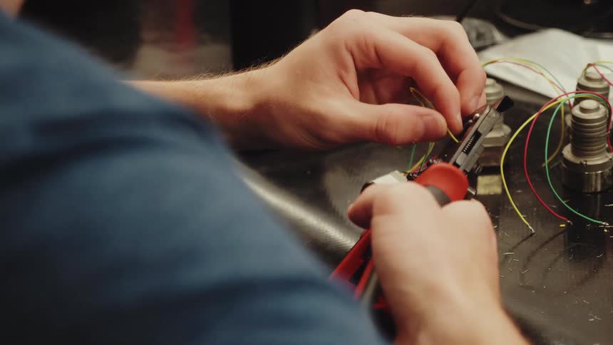 A persons hands wearing a blue shirt manipulate a wire stripping tool to remove insulation from a thin yellow electrical wire while other multicolored wires are connected to small metal parts on a dark workbench