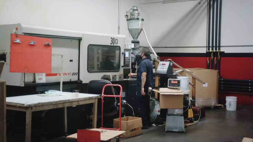 A worker wearing dark clothes and a mask operates a large injection molding machine in a factory setting with cement floors and red and black accent walls
