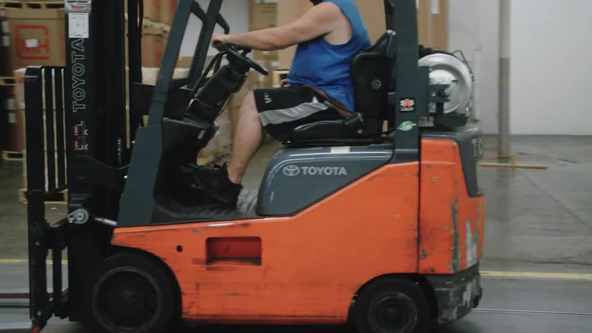 A close view shows a male worker wearing a blue shirt shorts and a face mask driving an orange toyota forklift down a wide aisle lined with pallets of cardboard boxes in a warehouse