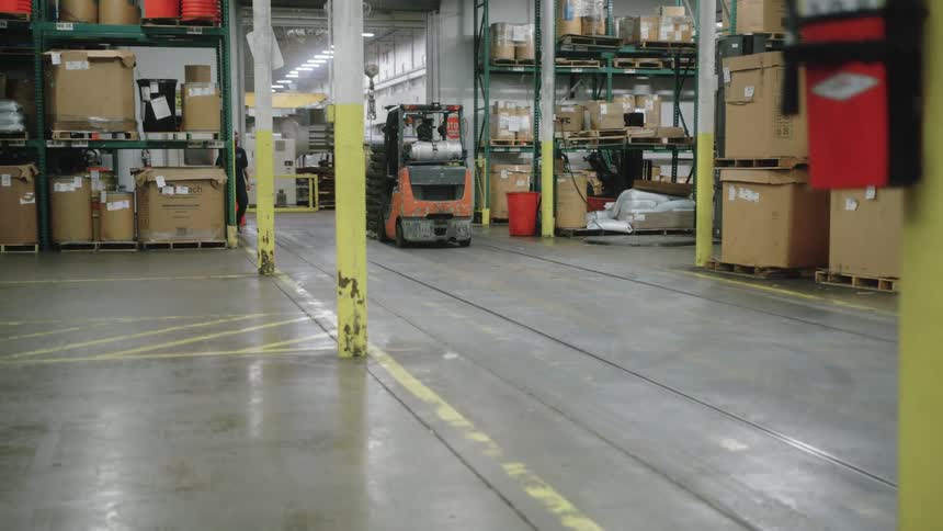 A medium view shows an orange forklift slowly backing up in a large warehouse aisle lined with tall shelves stacked high with various boxes and inventory