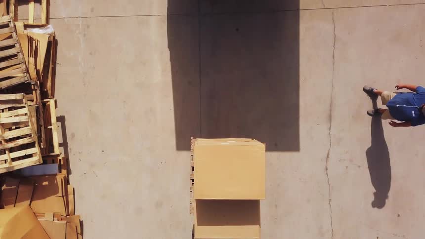 A power vehicle raises two cardboard containers out of a delivery truck reverses across a concrete lot past stacked wood platforms and lowers the containers toward the ground while a person watches