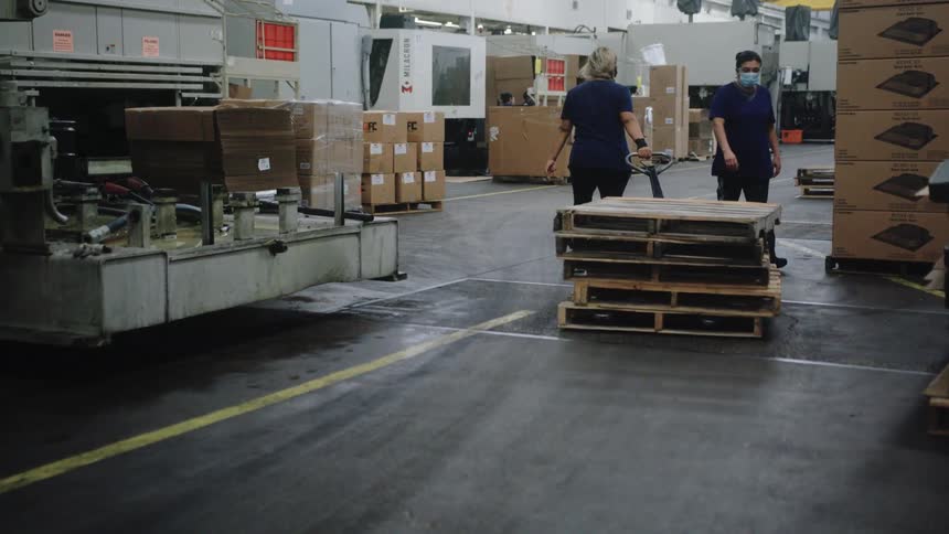 A long view of two female workers wearing dark blue uniforms and face masks as they walk with a pallet jack past stacks of wooden pallets and industrial machinery in a brightly lit factory