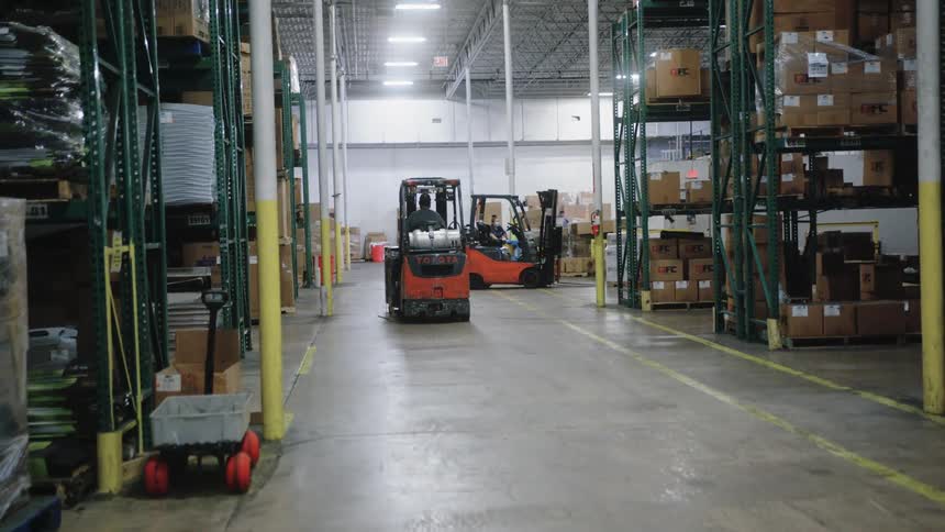 A forklift drives away down a long aisle in a brightly lit well stocked warehouse passing another forklift before parking at the far end
