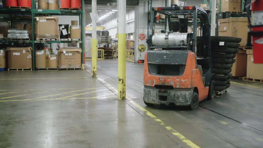 A person operates an orange vehicle carrying a stack of round objects the machine travels across a concrete floor with yellow markings and the background contains multiple rows of metal shelves holding various containers and boxes