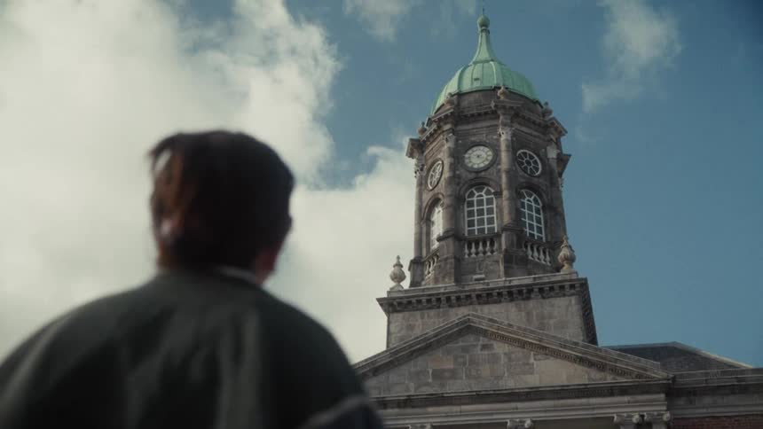 A low angle close view from behind a person shows the classic clock tower of dublin castle with a green cupola rising against a bright blue and cloudy sky