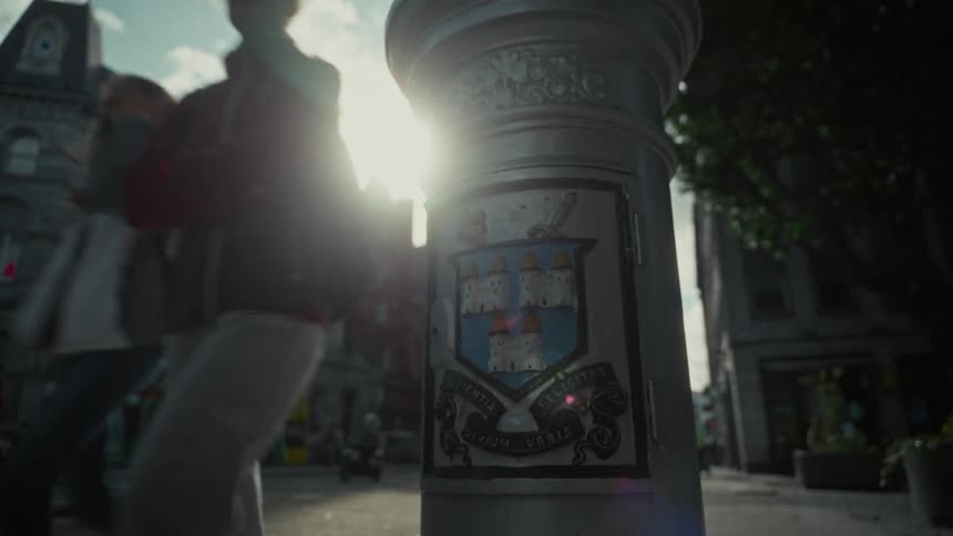 A camera shot focuses on the emblem on a metal street column while a lens flare from the sun obscures part of the view of the buildings people walking in the background and traffic on the streets