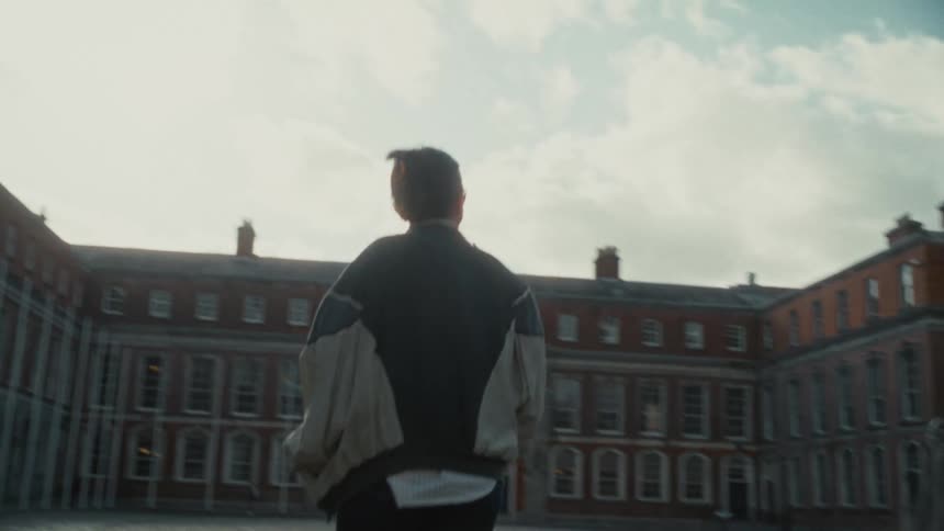 A shot from behind shows a person in a dark jacket with a white back panel walking across a paved courtyard towards a large classic brick building with multiple tall windows the architecture suggests a university campus or historic institution