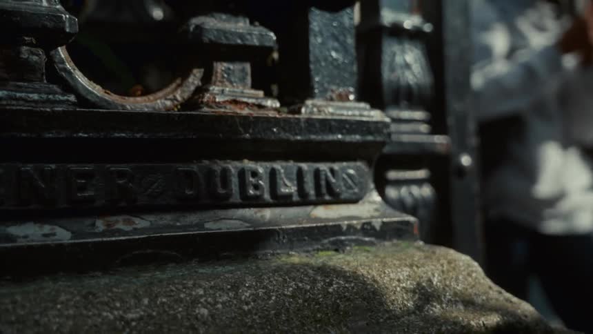 A close view of the corroded base of a dark metal railing shows the faded engraved text on a rough gray stone with a blurry person in the background