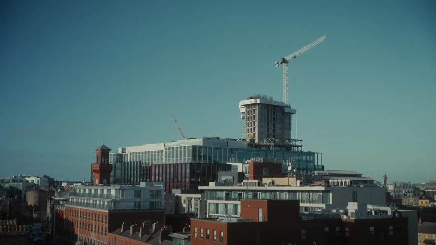 An urban landscape dominated by a building under construction on a hilltop featuring a tall tower crane and contrasting with the lower red brick buildings in the foreground against a clear sky