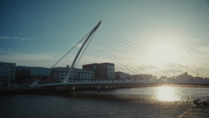 A modern bridge over water with sunlight reflecting brightly on the surface and surrounding buildings