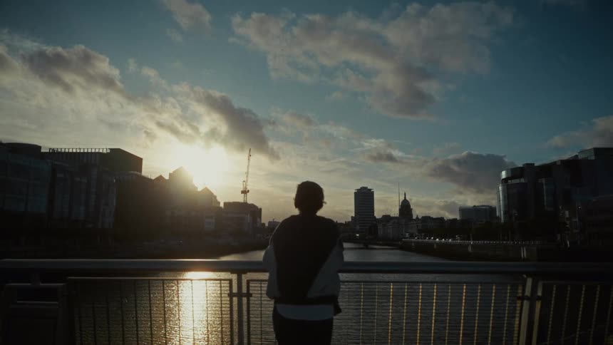 A woman leaning on a bridge railing over a river watching the city sunset
