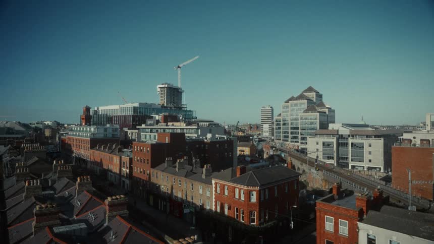 The video shows a highangle static shot of a cityscape featuring a mix of modern and traditional architecture under a clear blue skyin the foreground there are rows of older redbrick residential rooftops contrasting with the taller contempor