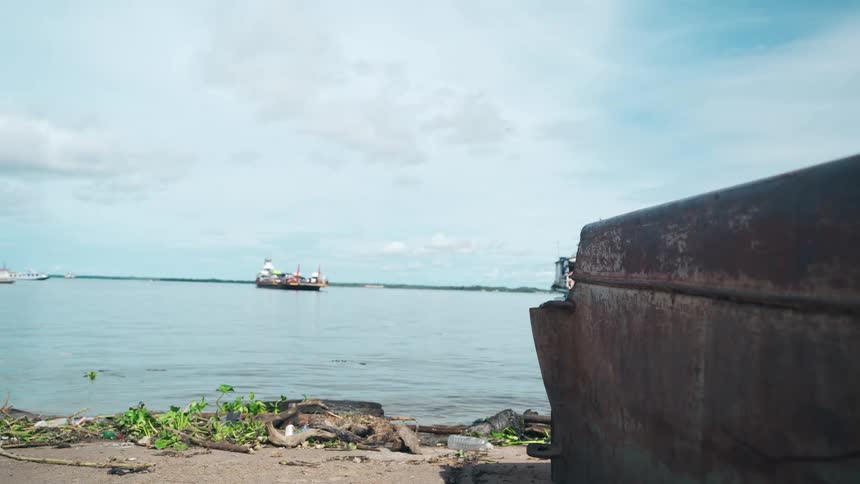 A cargo ship sails across a wide gray river under a cloudy sky seen from a muddy littered shoreline with an out of focus rusty metal object in the foreground