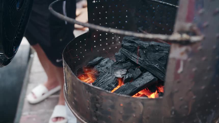 A closeup shot shows a person lighting up charcoal briquettes inside a metal charcoal chimney starter or small grillthe glowing red embers at the bottom are visible and the black briquettes are beginning to catch fire from below