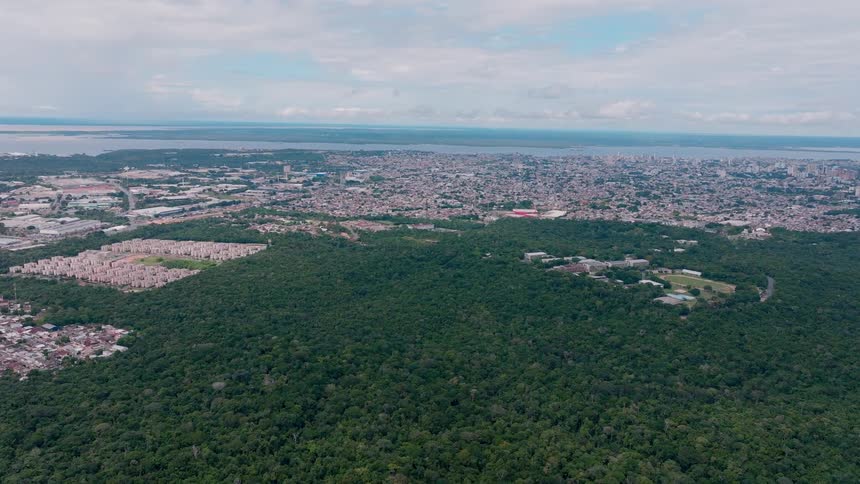 An expansive aerial view showcases the dense line where a large green forest meets a sprawling urban city which stretches out to the wide bright river on the horizon
