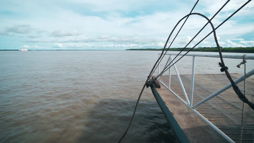 A broad brown river stretches to a distant shore under soft clouds with taut cables arching from a docked craft in the foreground