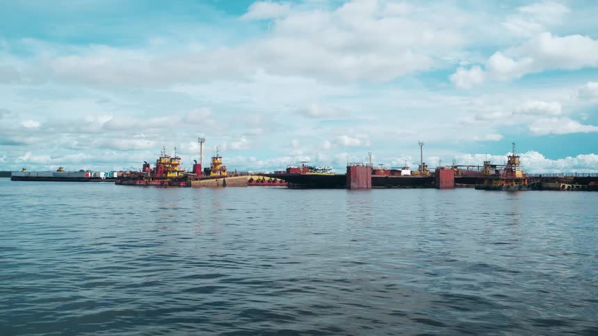 The video is a wide establishing shot of a large collection of brightly colored rusty tugboats and vessels moored together in a large body of waterthe ships dominate the midground against a light blue sky with scattered clouds suggesting a w