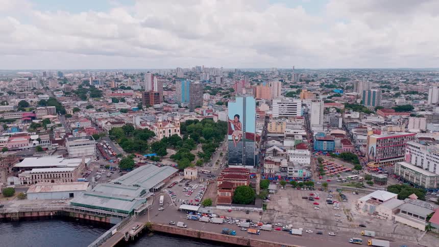 A camera shows a wide view of a densely constructed city numerous buildings of various sizes fill the land area a prominent tall structure displays a large figure image and a body of water and a bridge occupy the lower foreground