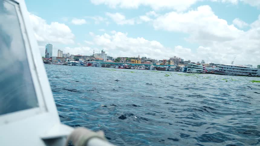 The skyline of a city with a mix of buildings and moored vessels is viewed from a low angle on a moving boat across a wide expanse of dark water under a partially cloudy sky