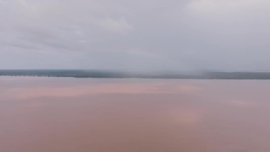 The video presents a wide aerial shot of a vast body of reddishbrown water likely a river or a large lake dominating the foreground the horizon features a dense dark green line of foliage suggesting a remote or heavily forested area all set