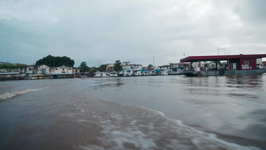 A camera moves across a wide expanse of brown water with surface waves toward a row of crowded hosues along a waterfront under an overcast sky with a red fuel dock structure visible on the right