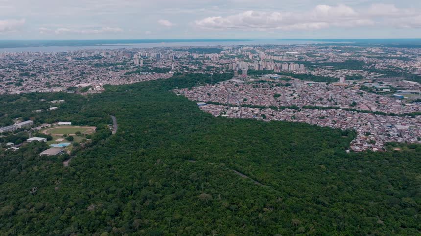 The video is an aerial wide shot showing a massive continuous expanse of dark green rainforestthe foreground of the forest is bordered by a densely populated city of lightcolored buildings that stretch to a distant horizon under an overca