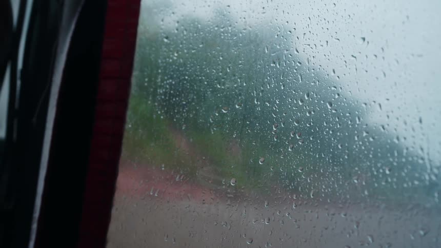 A camera shot focuses on a glass window covered in falling water droplets blurring the outoffocus green and gray landscape outside the vehicle
