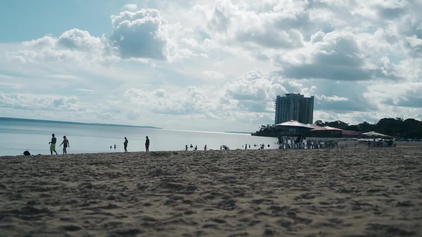 A wide low angle shot of a sandy beach shows silhouetted people walking along the waters edge with a tall highrise building and an overcast sky in the background